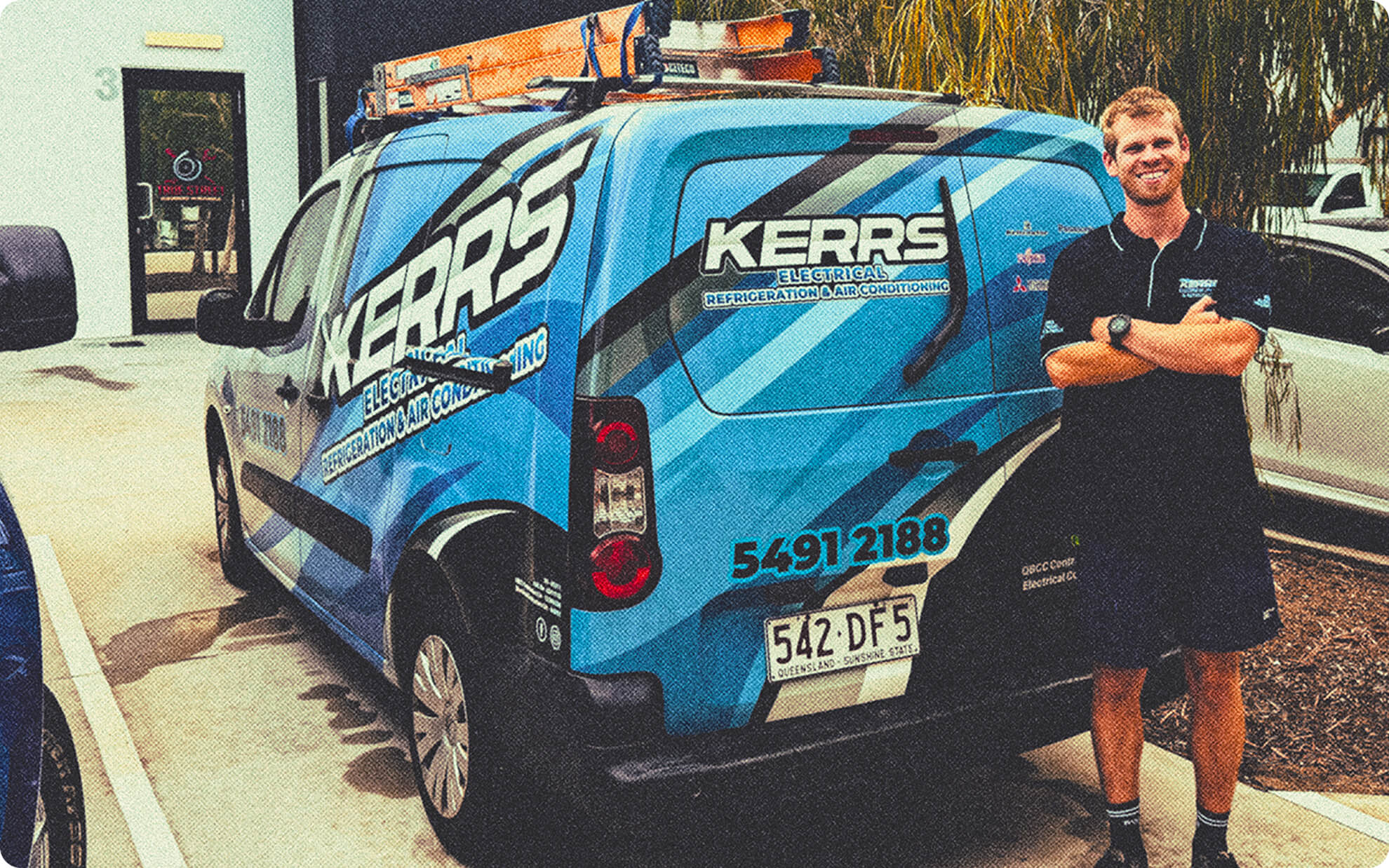 A man in a dark uniform stands smiling with arms crossed next to a blue Kerrs Electrical service van parked near a building.