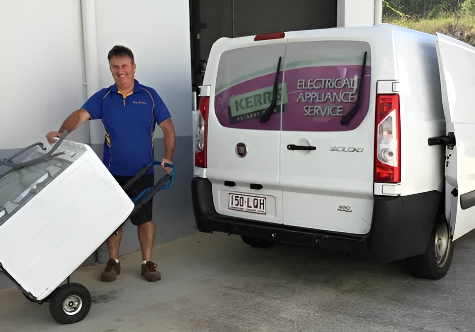 A man wearing a blue shirt moves an appliance on a hand truck toward a white service van labeled "Electrical Appliance Service" parked at a building.