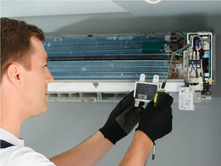 A person wearing gloves uses a digital multimeter to check the wiring of an open wall-mounted air conditioning unit.