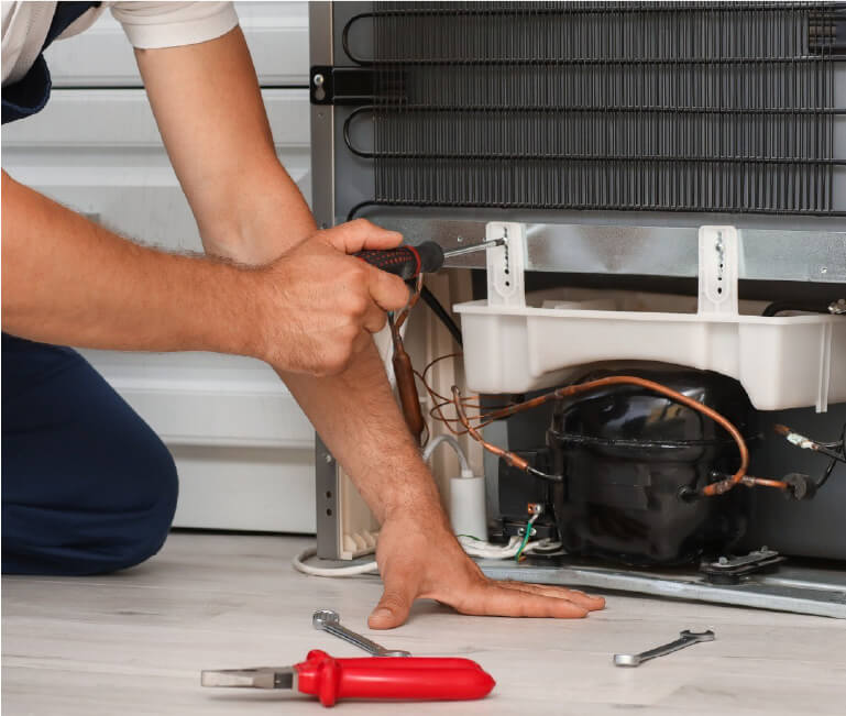 Person using a screwdriver to repair the lower rear section of a refrigerator, with various tools lying on the floor nearby.